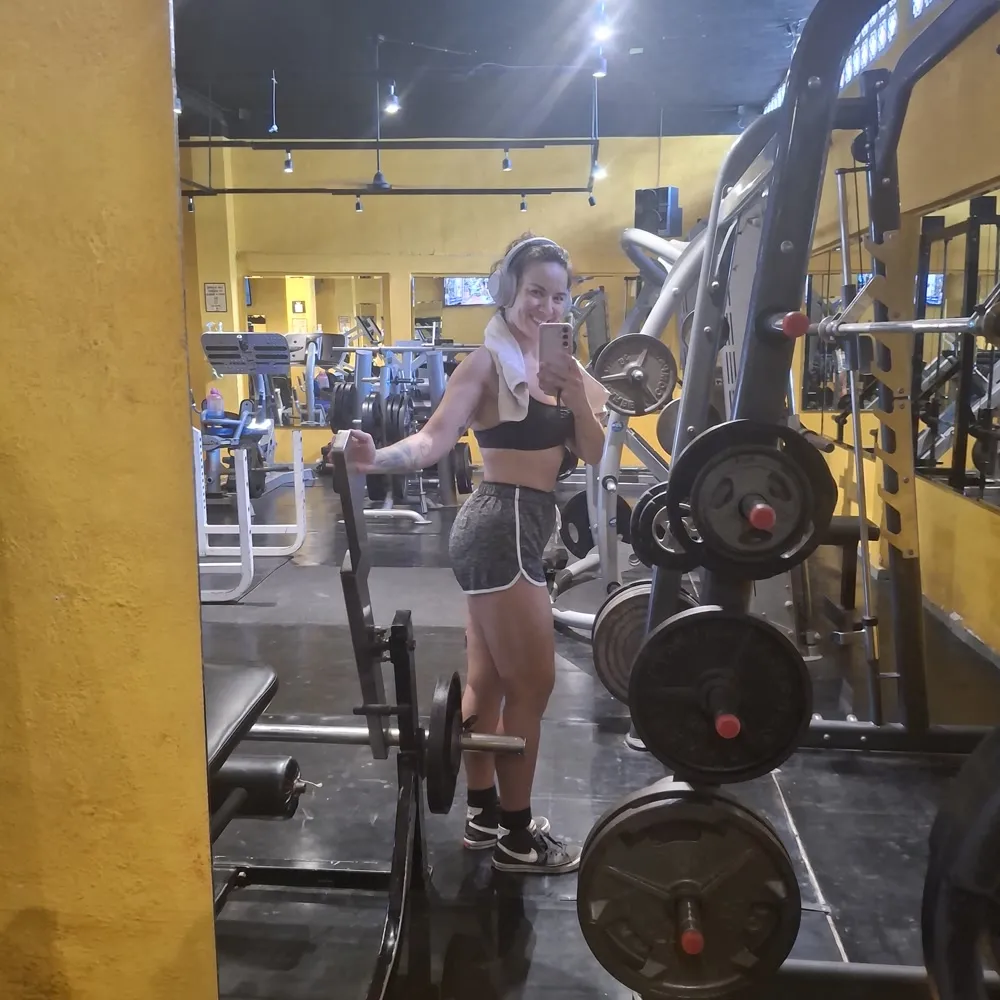 Gal Dorado in the gym mirror beside a loaded barbell and weight plates